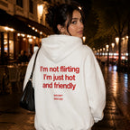 A woman standing on a city street at night wearing a white hoodie with red text on the back that reads 'I'm not flirting I'm just hot and friendly' and smaller text below, paired with black pants and carrying a black shoulder bag.