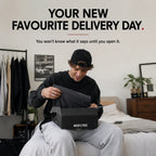 A man sitting on a bed in a bedroom opens a black NOFLTRS shoe box, revealing a dark gray shoe inside. Large promotional text at the top reads 'YOUR NEW FAVOURITE DELIVERY DAY.